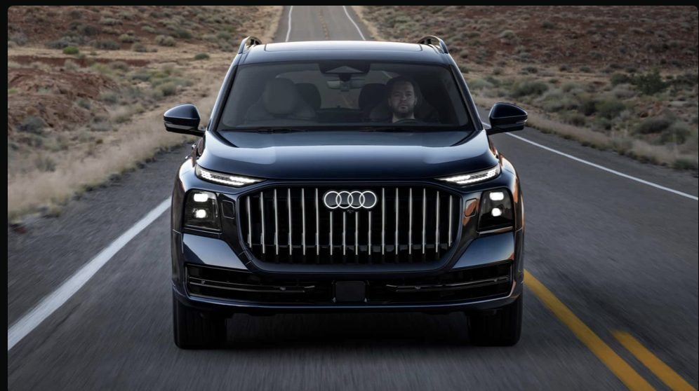 2027 Audi Q9 front-on view on a desert highway, large vertical-slat grille centered, dark navy exterior