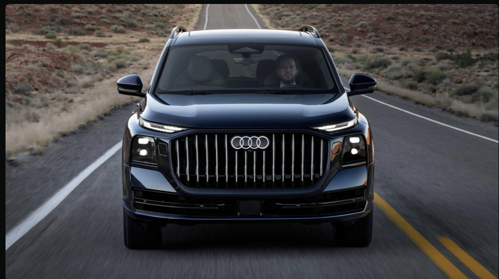 2027 Audi Q9 front-on view on a desert highway, large vertical-slat grille centered, dark navy exterior